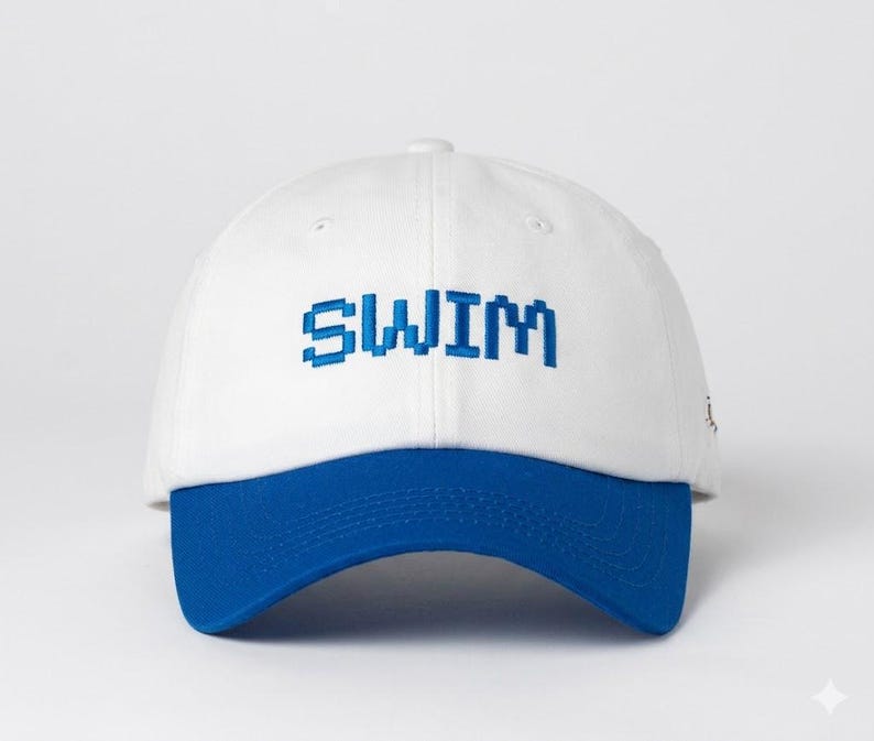 May include: A white baseball cap with a structured crown and a solid blue brim. The word SWIM is embroidered across the front center in a blue, pixelated, block-style font. The cap features a button on top and stitched eyelets for ventilation.