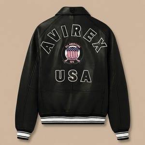 K&ouml;nnte beinhalten: Eine schwarze Lederjacke mit dem wei&szlig;en Schriftzug AVIREX im oberen R&uuml;ckenbereich, einem runden Aufn&auml;her mit amerikanischer Flagge und dem Text ALL AMERICANS ESTABLISHED 1975 sowie dem wei&szlig;en Schriftzug USA darunter, mit schwarz-wei&szlig; gestreiftem Rippstrick.