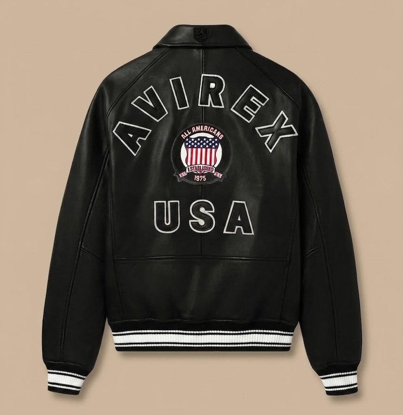 K&ouml;nnte beinhalten: Eine schwarze Lederjacke mit dem wei&szlig;en Schriftzug AVIREX im oberen R&uuml;ckenbereich, einem runden Aufn&auml;her mit amerikanischer Flagge und dem Text ALL AMERICANS ESTABLISHED 1975 sowie dem wei&szlig;en Schriftzug USA darunter, mit schwarz-wei&szlig; gestreiftem Rippstrick.