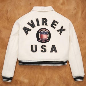Puede incluir: Chaqueta bomber color crema con ribete a rayas blancas y negras. La parte trasera presenta la palabra "AVIREX" sobre un emblema circular con la bandera estadounidense y el texto "USA". La chaqueta tiene un cuello cl&aacute;sico.