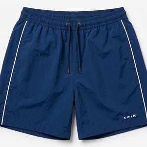 May include: Navy blue swim shorts featuring a white piped trim along the sides, an elasticized waistband with a matching navy blue drawstring, and the word SWIM printed in small white capital letters on the lower right leg area.