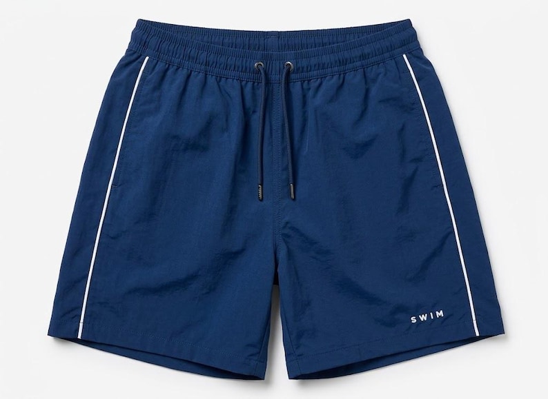 May include: Navy blue swim shorts featuring a white piped trim along the sides, an elasticized waistband with a matching navy blue drawstring, and the word SWIM printed in small white capital letters on the lower right leg area.