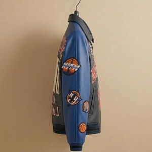 May include: A black and blue varsity jacket with basketball-themed patches. The jacket has a blue collar and sleeves, with patches featuring basketballs and the text "NY Basketball". The jacket is hanging on a black hanger.