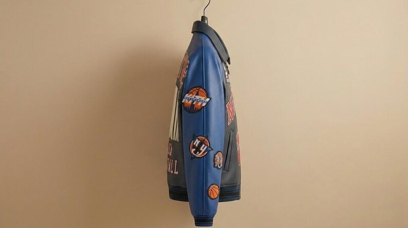 May include: A black and blue varsity jacket with basketball-themed patches. The jacket has a blue collar and sleeves, with patches featuring basketballs and the text "NY Basketball". The jacket is hanging on a black hanger.