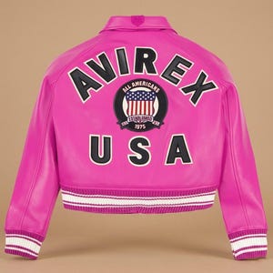 Puede incluir: Chaqueta de cuero rosa brillante con la palabra "AVIREX" en letras negras y blancas en la espalda. Debajo de la palabra "AVIREX" hay un parche circular con "ALL AMERICANS" y "ESTABLISHED 1975". La palabra "USA" est&aacute; debajo. La chaqueta tiene pu&ntilde;os y dobladillo a rayas.