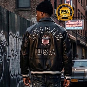 K&ouml;nnte beinhalten: Eine schwarze Lederjacke mit dem wei&szlig;en Schriftzug AVIREX USA auf dem R&uuml;cken sowie einem runden Aufn&auml;her mit einer amerikanischen Flagge. Die Jacke hat einen Kragen, lange &Auml;rmel mit gestreiften Rippb&uuml;ndchen und einen passenden gerippten Saum.