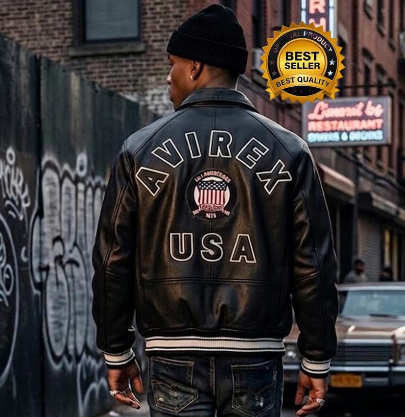K&ouml;nnte beinhalten: Eine schwarze Lederjacke mit dem wei&szlig;en Schriftzug AVIREX USA auf dem R&uuml;cken sowie einem runden Aufn&auml;her mit einer amerikanischen Flagge. Die Jacke hat einen Kragen, lange &Auml;rmel mit gestreiften Rippb&uuml;ndchen und einen passenden gerippten Saum.