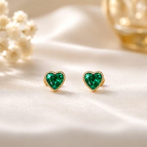 Peut inclure: Une paire de boucles d'oreilles à clous avec des pierres précieuses vertes facettées en forme de cœur, serties dans un métal doré poli. Chaque boucle présente une pierre vert émeraude avec une base pointue et un sommet arrondi.