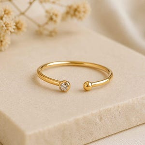 Peut inclure: Un bracelet manchette ouvert de couleur or. Une petite pierre précieuse transparente orne une extrémité, et une petite boule dorée l'autre. Le bracelet repose sur une surface beige clair, avec un élément floral flou en arrière-plan.