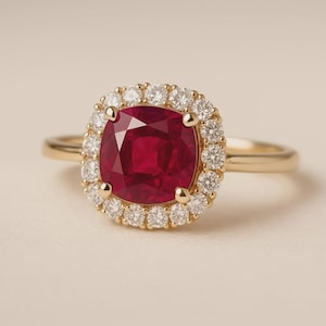 May include: A gold ring featuring a large, cushion-cut, deep red ruby at the centre, surrounded by a halo of small, round, brilliant-cut white diamonds. The band is crafted from polished yellow gold, providing a classic and elegant appearance.
