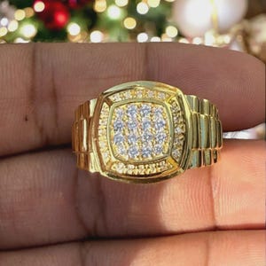 Könnte beinhalten: Ein goldfarbener Ring mit einer quadratischen Fassung in der Mitte, die mit zahlreichen kleinen, klaren und funkelnden Steinen besetzt ist. Die Ringschiene ist mit einem strukturierten Gliedermuster versehen, das an ein Metalluhrenarmband erinnert.
