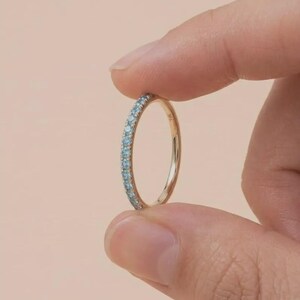 Puede incluir: Un anillo de banda de oro con una fila de pequeñas piedras preciosas de color azul claro. El anillo se sostiene entre dos dedos sobre un fondo rosa suave. El anillo parece ser de talla 14.