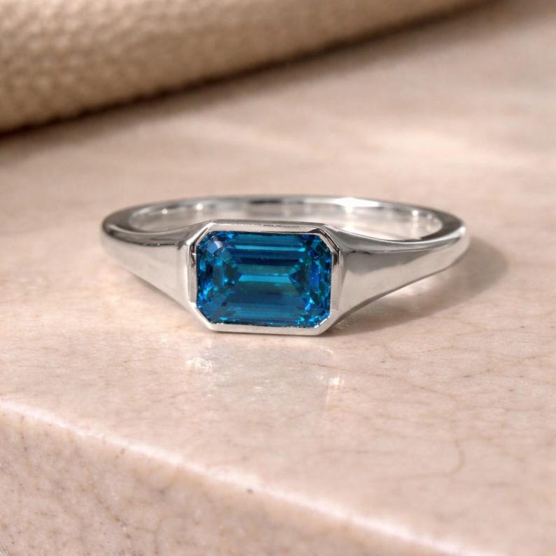 Op de afbeelding: Een zilveren ring met een rechthoekige, smaragdgeslepen, helderblauwe edelsteen. De ring heeft een gladde, gepolijste band en een bezel setting. De ring wordt gepresenteerd op een lichtgekleurd oppervlak.