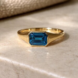 Op de afbeelding: Een gouden ring met een rechthoekige, smaragdgeslepen, blauwe edelsteen. De ring heeft een gladde, gepolijste band en een bezel setting. De ring wordt gepresenteerd op een lichtgekleurd oppervlak.