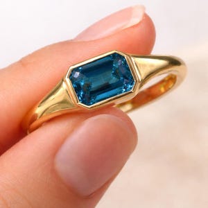 Op de afbeelding: Een gouden ring met een rechthoekige, gefacetteerde, blauwgroene edelsteen. De ring heeft een eenvoudig, elegant ontwerp met een gladde, gepolijste band. De edelsteen is gezet in een bezel setting. Sieraden.