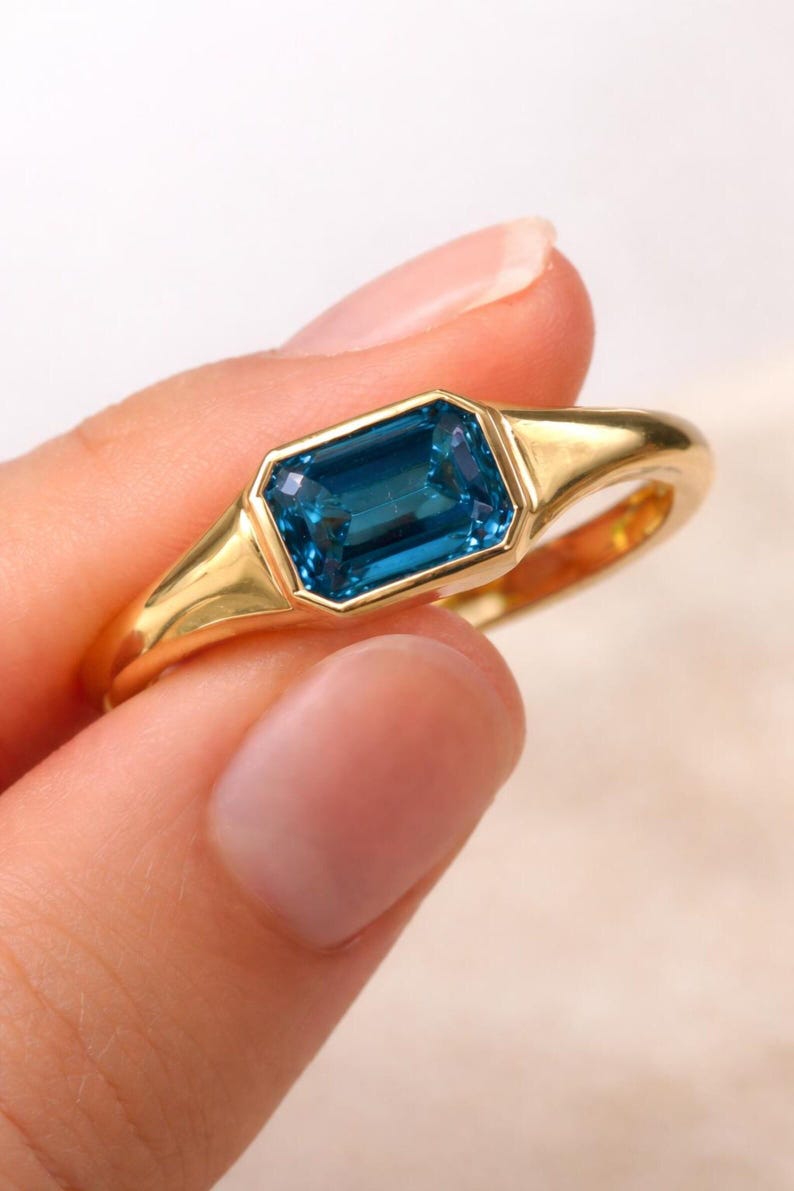 Op de afbeelding: Een gouden ring met een rechthoekige, gefacetteerde, blauwgroene edelsteen. De ring heeft een eenvoudig, elegant ontwerp met een gladde, gepolijste band. De edelsteen is gezet in een bezel setting. Sieraden.