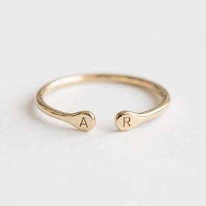 Puede incluir: Un anillo abierto de tono dorado con dos extremos redondeados y aplanados. Cada extremo está grabado con una letra mayúscula, con la letra A en el lado izquierdo y la letra R en el lado derecho. La banda es delgada y tiene un acabado pulido.