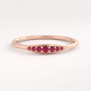 Op de afbeelding: Een roségouden ring met een rij van zeven kleine, ronde, rode edelstenen in een gebogen ontwerp. De ringband is glad en gepolijst, en de edelstenen zijn gelijkmatig verdeeld. De ring staat tegen een witte achtergrond.
