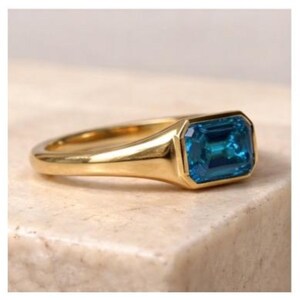Op de afbeelding: Een gouden ring met een rechthoekige, smaragdgeslepen, teal-kleurige edelsteen. De ring heeft een gladde, gepolijste band en een bezel-zetting. De ring wordt gepresenteerd op een neutraal getinte ondergrond.