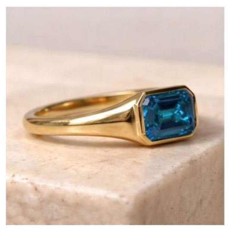 Op de afbeelding: Een gouden ring met een rechthoekige, smaragdgeslepen, teal-kleurige edelsteen. De ring heeft een gladde, gepolijste band en een bezel-zetting. De ring wordt gepresenteerd op een neutraal getinte ondergrond.