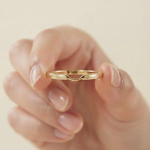 May include: A polished gold band ring featuring a subtle, curved dip along the top edge. The ring has a smooth, rounded profile and a high-shine metallic finish, designed with a contoured shape that allows it to sit flush against another ring.