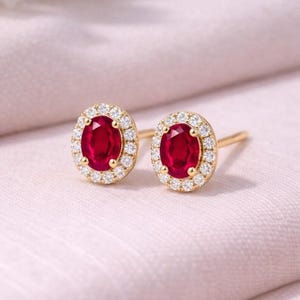 Peut inclure: Une paire de boucles d'oreilles à clous en or, chacune ornée d'une pierre précieuse ovale rouge vif entourée d'un halo de petits diamants blancs scintillants. Les boucles d'oreilles sont posées sur un fond rose tendre.