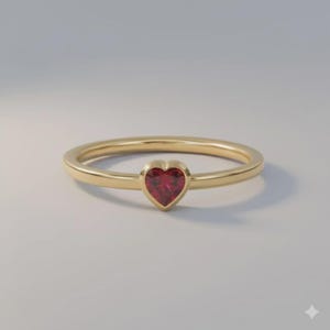 May include: A gold ring featuring a single, small, heart-shaped red gemstone set in a bezel setting. The band is thin, smooth, and polished, creating a simple and clean design. The red stone is centred on the band, providing a focal point.