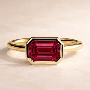 Puede incluir: Un anillo de oro con una piedra preciosa rectangular de color rojo intenso. La piedra preciosa está engastada en un bisel de oro, y la banda es de oro liso y pulido. El anillo se muestra sobre una superficie clara.