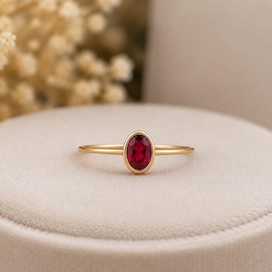 Puede incluir: Un anillo de oro con una vibrante gema roja ovalada. El anillo se presenta sobre una superficie de color neutro, con elementos de fondo suaves y borrosos. La delgada banda del anillo resalta la prominente gema.