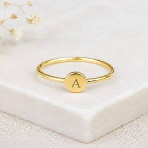 Op de afbeelding: Een goudkleurige ring met een dunne band en een cirkelvormige bedel met de letter "A". De ring wordt gepresenteerd op een wit, getextureerd oppervlak. Op de achtergrond een takje met kleine, witte bloemen. Het ontwerp van de ring is minimalistisch.