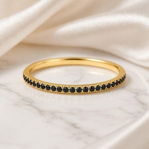May include: A gold-colored ring with a band of small, black gemstones. The ring is set against a white marble surface and a backdrop of draped, off-white fabric. The ring's design is simple and elegant.