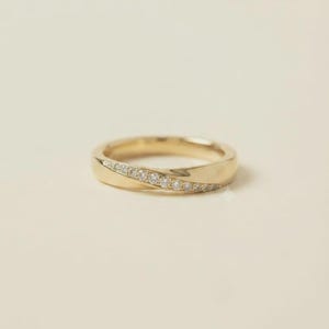May include: A gold ring featuring a polished band with a diagonal channel set with a row of small, clear, round-cut stones. The metal has a smooth, reflective finish, and the stones are arranged in a single line across the center of the band.