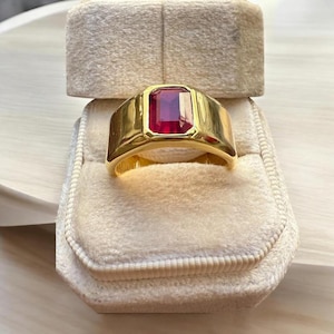May include: A gold ring with a rectangular, deep red gemstone. The ring is set in a thick, polished gold band. The ring is displayed in a beige velvet ring box.