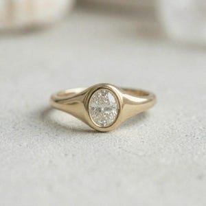Könnte beinhalten: Ein goldener Siegelring mit einer glatten, polierten Ringschiene und einem zentralen, ovalen, klaren Edelstein in einer Zargenfassung. Der Ring hat ein massives, abgerundetes Profil mit einer Schiene, die sich zur Mitte hin verbreitert.