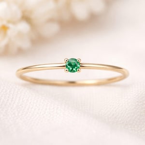 Può includere: Un delicato anello d'oro con una piccola e rotonda gemma di smeraldo verde brillante. L'anello ha una sottile fascia in oro lucido. Lo smeraldo è il punto focale, incastonato in una semplice montatura a quattro griffe.
