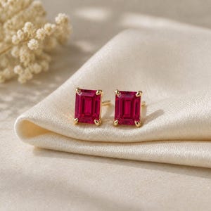 Peut inclure: Une paire de boucles d'oreilles à clous en or avec des pierres précieuses rectangulaires rouge foncé. Les boucles d'oreilles sont posées sur un tissu de satin crème, avec une mise au point douce en arrière-plan.