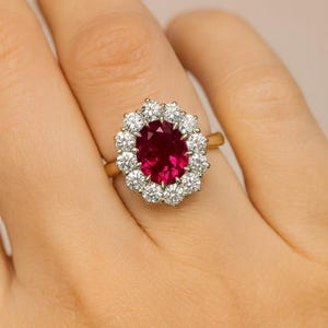 Op de afbeelding: Een gouden ring met een centrale ovaal geslepen dieprode robijn, omringd door een halo van twaalf ronde, briljant geslepen witte diamanten. De edelstenen zijn gezet in een klauwzetting op een gepolijste gouden band rond de vinger.