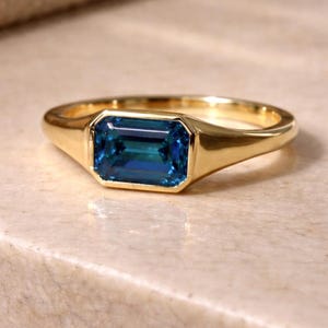 May include: A gold ring with a rectangular, emerald-cut, teal-colored gemstone. The ring has a smooth, polished band and a bezel setting that securely holds the gemstone. The ring is displayed on a light-colored surface.