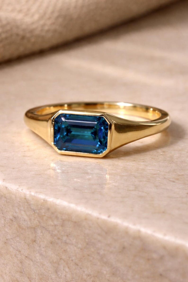 Op de afbeelding: Een gouden ring met een rechthoekige, smaragdgeslepen, teal-kleurige edelsteen. De ring heeft een gladde, gepolijste band en een bezel setting. De ring wordt gepresenteerd op een lichtgekleurd oppervlak.