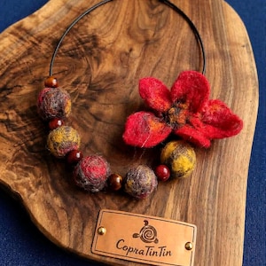 Può includere: Una collana fatta a mano con un vibrante fiore rosso in feltro e una serie di perline di lana infeltrita nei toni del rosso, giallo e marrone. La collana è esposta su una superficie di legno e include un'etichetta in pelle con il testo "CopraTinTin".