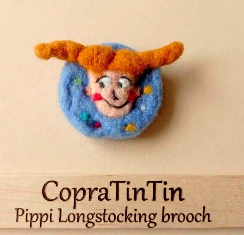 Puede incluir: Broche hecho a mano con una cara de dibujos animados con pelo naranja y fondo azul. El broche tiene el texto "CopraTinTin Pippi Longstocking brooch" en una superficie de madera. La cara tiene mejillas rojas y una expresi&oacute;n sonriente.