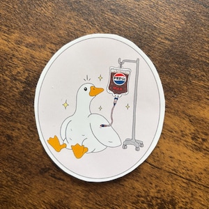 May include: A circular sticker showing a cartoon duck receiving an IV drip of Pepsi Max. The duck is white with an orange beak and feet, and the IV bag has the Pepsi logo. The sticker has a light pink border and is on a brown surface.