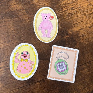 Peut inclure: Trois autocollants sur une surface en bois. L'un représente un ours en peluche rose avec une étiquette en forme de cœur rouge. Un autre montre une créature rose avec un nœud papillon jaune. Le troisième est un animal de compagnie numérique rose et vert.