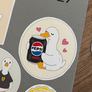 May include: A circular sticker featuring a white duck with an orange beak and feet, holding a black Pepsi Max soda can. The duck has its eyes closed and is surrounded by three small pink hearts against a light cream-coloured circular background.