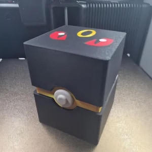 以下が含まれることがあります： マットブラックの正方形の収納ボックス。上蓋には、様式化された目と円形のエンブレムを模した黄色と赤のグラフィックデザインが施されています。ボックスの中央には、突き出た白い円形のボタンが付いた水平の黄色い帯があります。