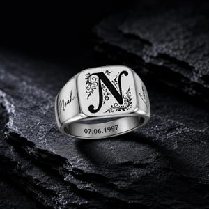 May include: A silver-toned signet ring featuring a large engraved letter N with decorative floral flourishes on the face. The sides of the ring are engraved with the names Noah and Liam, while the band displays the date 07.06.1997 in black text.