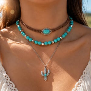 Puede incluir: Un conjunto de collares superpuestos que incluye una gargantilla de cordón marrón con una piedra turquesa ovalada, una hilera de cuentas turquesas con espaciadores plateados y una cadena plateada con un colgante de cactus texturizado.