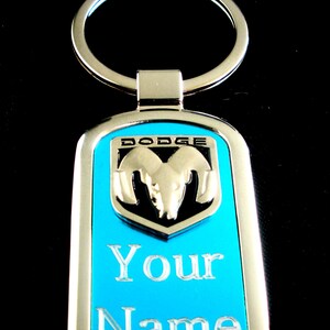 May include: A rectangular metal keychain with a bright blue center panel. It features a silver-toned Dodge ram logo at the top and the text "Your Name" engraved in silver lettering below the logo. The keychain includes a circular silver split ring.