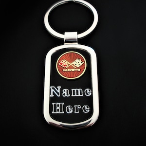 Puede incluir: Un llavero rectangular de metal plateado con un centro negro. El llavero presenta el logotipo de Corvette en rojo y dorado, con el texto "Name Here" en blanco. Un llavero plateado está adjunto.