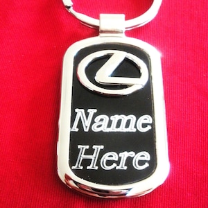 Puede incluir: Un llavero rectangular de metal con un borde plateado pulido y un centro negro. La parte superior muestra un logotipo de Lexus plateado en relieve, mientras que la parte inferior está grabada con el texto blanco Name Here en fuente serif.